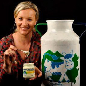 Anne Lenormand tenant un pot de crème fraîche à côté d'un bidon de lait peint avec le logo historique